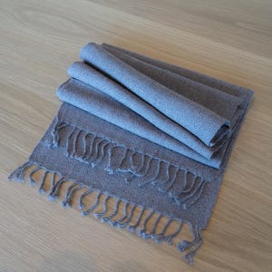 May include: A folded, gray scarf with a fringed edge. The scarf is made of a soft, woven material and is displayed on a light-colored wooden surface. The scarf's color is a muted gray-blue.