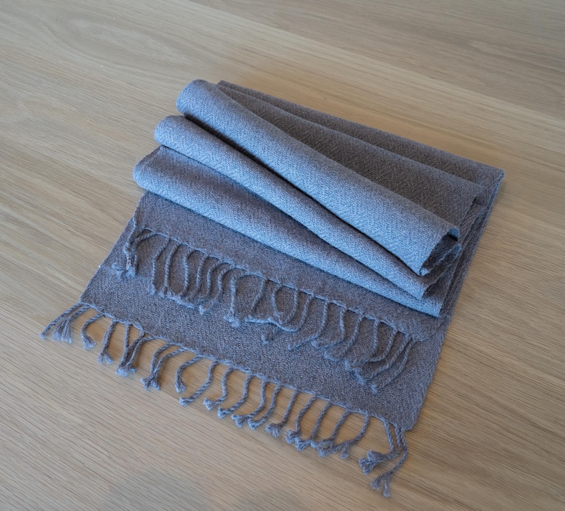 May include: A folded, gray scarf with a fringed edge. The scarf is made of a soft, woven material and is displayed on a light-colored wooden surface. The scarf's color is a muted gray-blue.