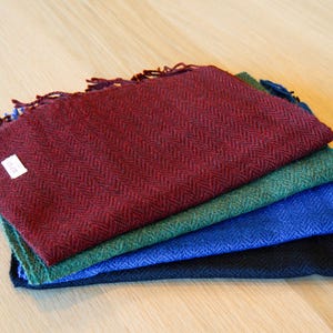 May include: A stack of four woven scarves in shades of burgundy, green, blue, and black. Each scarf has a herringbone pattern and fringed edges. A small white tag is visible on the burgundy scarf.