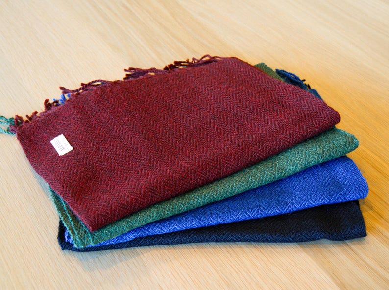 May include: A stack of four woven scarves in shades of burgundy, green, blue, and black. Each scarf has a herringbone pattern and fringed edges. A small white tag is visible on the burgundy scarf.