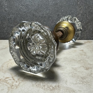 May include: A pair of antique glass doorknobs. The knobs are clear with a faceted design, and the hardware is brass and bronze. The doorknobs are resting on a light-colored surface against a dark gray background.
