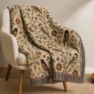 May include: A woven throw blanket featuring a cream-colored background with a repeating pattern of various birds, red flowers, and green leafy vines. The blanket has a textured appearance and includes a decorative fringe along the bottom edge.