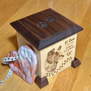 May include: Wooden pet urn with a dark brown lid and paw print design. The urn features a laser-etched image of a dog and the name "El Bee". A heart-shaped memorial with a ribbon is also present.