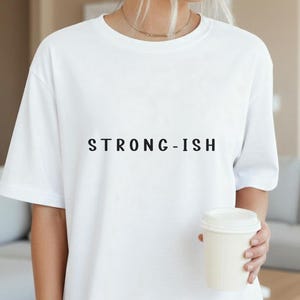 Könnte beinhalten: Weißes T-Shirt mit dem Text "STRONG-ISH" in Schwarz. Das Shirt hat einen Rundhalsausschnitt. Das Wort "White" ist unten links im Bild aufgedruckt. Eine Person hält einen weißen Kaffeebecher.