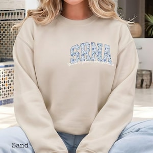 May include: Sand-colored sweatshirt with the letters "SRNA" in a floral pattern across the chest. Below the letters is the text "Student Registered Nurse Anesthetist". The sweatshirt has long sleeves and a crew neck.