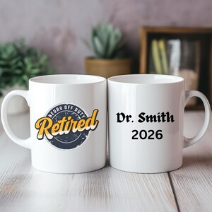 May include: Two white ceramic mugs. One mug features the text "Retired" in yellow script with a navy blue and distressed design, and the words "Neuro Off Duty". The other mug has "Dr. Smith 2026" in black text.