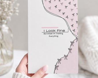 May include: A rectangular notebook with a white cover featuring a soft pink section on the right side decorated with small black bow icons. The text I Look Fine But Inside I'm Tracking Everything is printed in black with a pink horizontal line above.