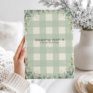 May include: A rectangular notebook with a light sage green and cream checkered pattern. The cover features the text "Dealing With It" and "A No-Nonsense Daily" in black font, surrounded by watercolour-style eucalyptus leaf illustrations at the corners.