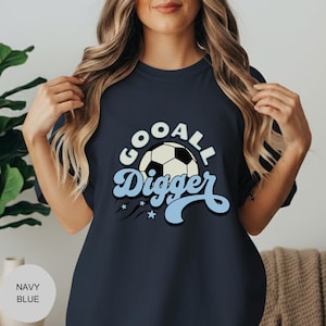 May include: Navy blue t-shirt with the text "GOOALL Digger" in a retro font, featuring a soccer ball graphic. The design includes light blue accents and stars. The shirt is a casual wear item.