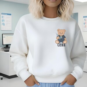 May include: A white sweatshirt featuring a cartoon teddy bear wearing a nurse's uniform and holding a syringe. The text "CRNA" is printed below the bear. The sweatshirt is a solid white color.