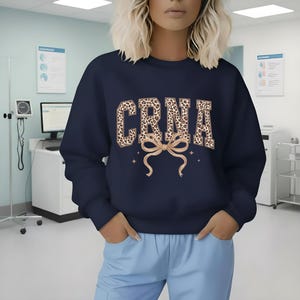May include: Navy blue sweatshirt featuring the letters "CRNA" in a leopard print pattern, a tan bow, and star details. The sweatshirt is worn with light blue trousers.