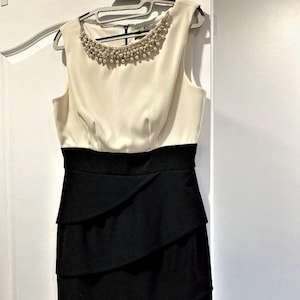 May include: Sleeveless ivory and black dress with a pearl-beaded neckline. The bodice is ivory, transitioning to a black, layered skirt. The dress hangs on a white hanger against a white door.