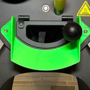 Könnte beinhalten: Nahaufnahme eines schwarzen und grünen Kaffeerösters. Der Röster hat eine leuchtend grüne Tür und Akzente, einen schwarzen Griff und ein gelbes Warnsymbol. Der Röster steht vor einem neutralen Fliesen Hintergrund.