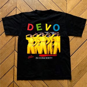 May include: Black t-shirt featuring the band name "DEVO" in colorful letters above a graphic of the band members in yellow jumpsuits. The text "FOUR SHOWS!! IN CONCERT!!" is printed below the graphic.
