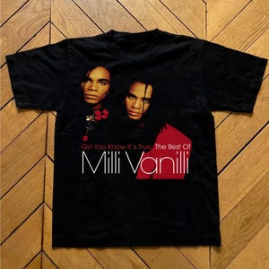 May include: Black t-shirt featuring a graphic of two people with long hair and the text "Girl You Know It's True The Best Of Milli Vanilli". The design includes a red accent.