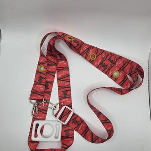 May include: Red lanyard with a repeating black pattern and the text "JIN". The lanyard has a white plastic clasp, metal swivel hooks, and gold-colored eyelets. The lanyard is designed to be worn around the neck.