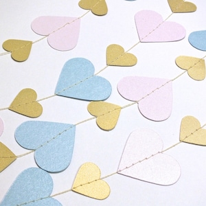 May include: A garland of paper hearts in pink, blue, and gold. The hearts are strung together with twine and are perfect for a party or wedding decoration.