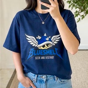 Puede incluir: Camiseta azul marino con un gr&aacute;fico de una concha azul con alas blancas y el texto "BLUESHELL SEEK AND DESTROY". La camiseta est&aacute; hecha de un material suave y tiene mangas cortas.