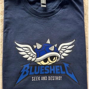 Puede incluir: Camiseta azul marino con un gr&aacute;fico de una concha azul con alas y el texto "BLUESHELL SEEK AND DESTROY". El dise&ntilde;o es en blanco y azul. La camiseta est&aacute; doblada sobre una superficie beige.