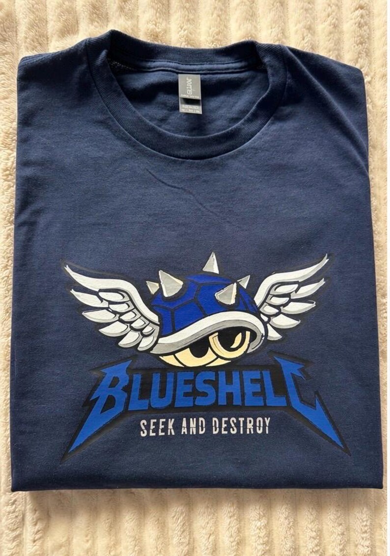 Puede incluir: Camiseta azul marino con un gr&aacute;fico de una concha azul con alas y el texto "BLUESHELL SEEK AND DESTROY". El dise&ntilde;o es en blanco y azul. La camiseta est&aacute; doblada sobre una superficie beige.