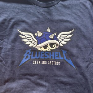 Puede incluir: Camiseta azul marino con un dise&ntilde;o de caparaz&oacute;n azul con alas blancas y el texto "BLUESHELL SEEK AND DESTROY" en una fuente estilizada. El dise&ntilde;o est&aacute; centrado en la parte delantera de la camiseta.