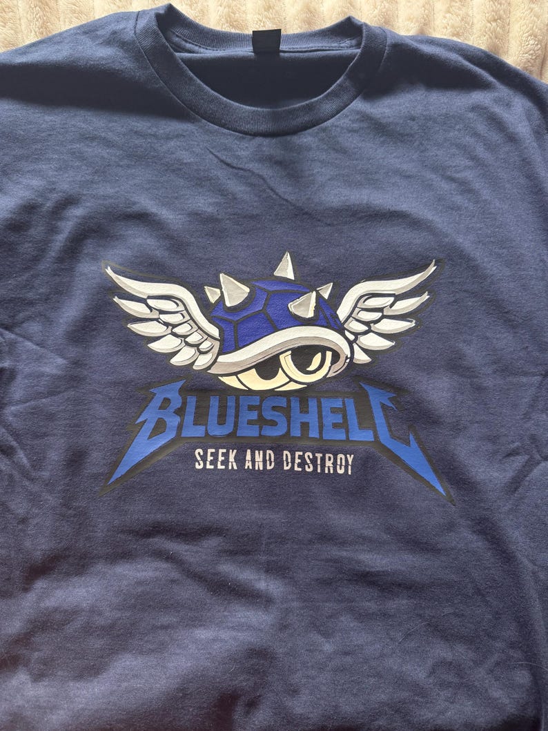 Puede incluir: Camiseta azul marino con un dise&ntilde;o de caparaz&oacute;n azul con alas blancas y el texto "BLUESHELL SEEK AND DESTROY" en una fuente estilizada. El dise&ntilde;o est&aacute; centrado en la parte delantera de la camiseta.