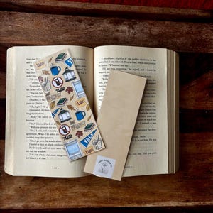 Könnte beinhalten: Ein offenes Buch mit einem dekorativen Aufkleberbogen und einem Holzlesezeichen. Der Aufkleberbogen zeigt verschiedene Illustrationen, darunter eine Kaffeetasse, eine Straßenbahn und das Wort "Yale". Das Lesezeichen trägt den Text "Poppy Meadow Co."