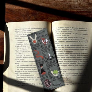 May include: A bookmark with illustrated squares featuring images and text. The bookmark is grey with black and red accents. The bookmark is on an open book with text.