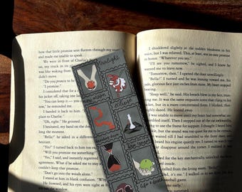 May include: A bookmark with a series of illustrated squares featuring images and text. The bookmark is gray with black and red accents. The bookmark is lying on top of an open book with text.
