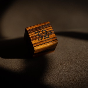 May include: A wooden die with the number 32 etched in black. The die is a cube shape with a striped pattern of light and dark brown wood. The die is set against a dark, textured background.