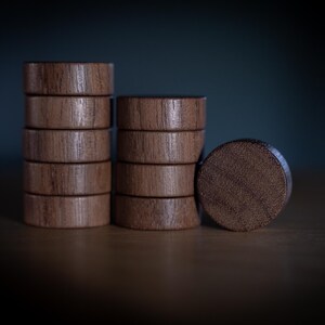 May include: A set of eleven circular wooden game pieces with a visible natural grain pattern. Five pieces are stacked in a column on the left, four pieces are stacked in a column in the center, and two pieces are positioned upright on the right.