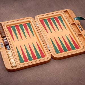 Könnte beinhalten: Ein Backgammon-Spielset aus Holz mit einem hellbraunen Rahmen, einer beigen Spielfläche, roten und grünen dreieckigen Punkten sowie zwei Sätzen runder Spielsteine aus hellem und dunklem Holz. Enthält Würfel, einen Verdopplungswürfel und einen grünen Griff.