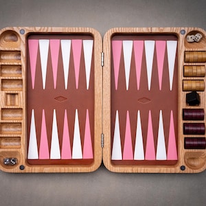 Könnte beinhalten: Ein hölzernes Backgammon-Spielbrett mit einer braunen Spielfläche und abwechselnd rosa und weißen dreieckigen Punkten. Das Brett verfügt über seitliche Aufbewahrungsfächer, die hölzerne Spielsteine in Hellbraun und Dunkellila sowie Würfel enthalten.