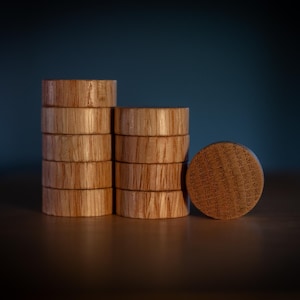May include: Wooden jar lids stacked in two columns and a single lid. The lids are made of light brown wood with a visible grain. The single lid has text etched into its surface. The background is a dark blue.