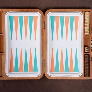Könnte beinhalten: Ein Backgammon-Spiel aus Holz mit einer weißen Spielfläche und abwechselnden türkisfarbenen und pfirsichfarbenen Dreiecken. Das Set enthält helle und dunkle Holzspielsteine, einen Verdopplungswürfel mit der Zahl 64 und einen grünen Ledergriff.