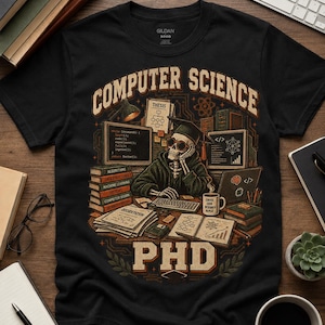 May include: A black t-shirt featuring a graphic of a skeleton wearing a graduation cap and glasses, sitting at a desk with computers, books, and papers. The text reads COMPUTER SCIENCE at the top and PHD at the bottom, with coffee and code.
