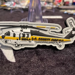 May include: A white, black, and yellow helicopter-shaped patch. The patch features the text "M921LA L.A. COUNTY FIRE" and the number "21". The helicopter has a black rotor and a yellow stripe along the bottom.