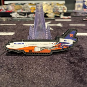 May include: A patch depicting a 10 Tanker aircraft, featuring a white fuselage with orange and blue accents. The patch includes the text "10 TANKER" and the number "914". The design is detailed with various elements.