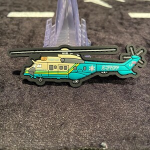 May include: A teal, tan, and black helicopter-shaped patch with the text "LOS ANGELES COUNTY SHERIFF" and "LB". The patch has a black border and is on a dark surface.