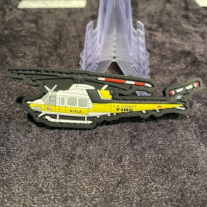 May include: A black, yellow, and white helicopter patch with the text "L.A. COUNTY FIRE" and "N14LA". The patch has a black border and a red and white striped tail.