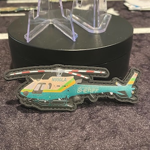 May include: A teal, yellow, and white helicopter patch with the text "N950LA" and "LOS ANGELES COUNTY SHERIFF". The patch has a black border and is displayed on a dark surface.