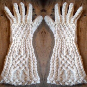 May include: A pair of cream-colored, knitted gloves with intricate cable and textured patterns. The gloves are long, extending past the wrist, and are displayed against a wooden background. The design features detailed stitching and a classic, warm aesthetic.