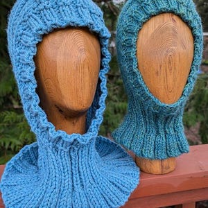 May include: Two knitted balaclavas in shades of blue. The balaclavas are displayed on wooden mannequin heads. The one on the left is a lighter blue, while the one on the right is a teal color. The knitted texture is visible, with ribbed detailing around the neck and face.