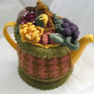 May include: A yellow teapot with a knitted cozy. The cozy features a floral design with yellow, purple, and green accents. The cozy is adorned with decorative beads and a scalloped edge. The teapot has a curved handle and spout.
