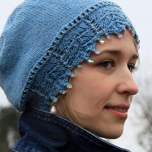 May include: A light blue knitted beret with a decorative lace trim and beaded accents. The beret is adorned with dangling iridescent beads. The hat is worn with a denim jacket.
