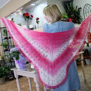 May include: A handmade, open-weave shawl in a gradient of pink and white. The shawl has a scalloped edge and a delicate, lacy pattern. The color transitions from a deep pink at the edges to white in the center. The shawl is held up, showcasing its full shape.