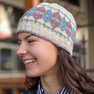 May include: A knitted beanie hat with a geometric pattern in red, blue, and green on a cream background. The hat has a ribbed brim and a contrasting trim. The hat is a winter accessory.