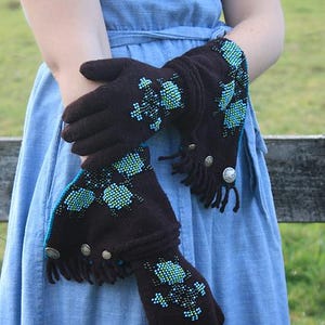 Könnte beinhalten: Dunkelbraune Handschuhe mit türkisfarbenen und hellblauen, perlenbesetzten Blumenmustern. Die Handschuhe haben Fransen und silberfarbene Knöpfe. Die Manschetten sind mit türkisfarbenem Besatz versehen. Die Handschuhe werden vor einem blauen Kleid präsentiert.
