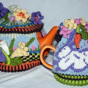 May include: Two ceramic teapots with orange handles and spouts, adorned with knitted covers. The covers are light blue, decorated with colorful knitted flowers and other floral elements. One teapot has a lid with a knitted flower arrangement.