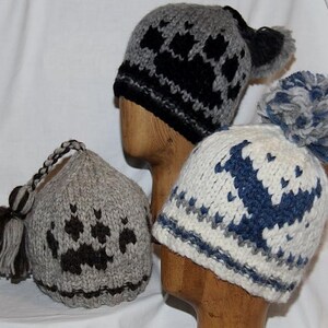 Può includere: Tre cappelli invernali lavorati a maglia in grigio, nero e bianco, ognuno con un design unico. Uno presenta un motivo a impronte di zampe, un altro ha un design geometrico e il terzo mostra un motivo blu e bianco. Ogni cappello è esposto su una testa di legno.
